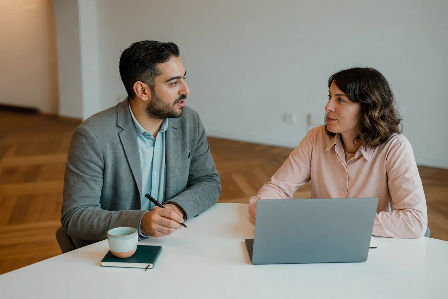 Two people talking across a table with a laptop and notebook.